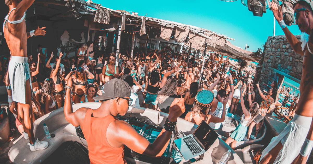 Luxury beach club party in Mykonos at sunset with guests dining and dancing beside the Aegean Sea.